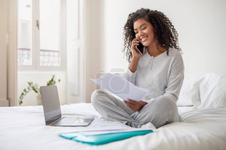 Una joven hispana con el pelo rizado sonríe mientras habla por teléfono, sostiene documentos en su mano, se viste con ropa cómoda y casual y está sentada en una cama con un portátil y carpetas cerca.
