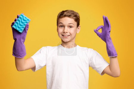 Foto de Concepto de limpieza. Lindo chico en guantes de limpieza que muestran OK signo y la celebración de la esponja, fondo panorama naranja - Imagen libre de derechos