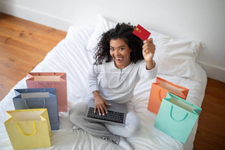 Vue de dessus de la femme hispanique est assis sur un lit, tenant une carte de crédit dans une main et un ordinateur portable dans l'autre. Elle semble être engagée dans des achats en ligne ou des transactions financières.