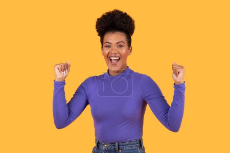 Hispanic woman wearing a purple shirt and jeans standing on yellow background, clenching fists. She is casually dressed in comfortable clothing