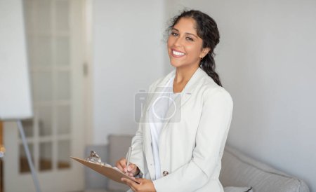Foto de Una mujer profesional con un traje blanco está de pie y sosteniendo un portapapeles. Ella aparece enfocada y comprometida en su trabajo. - Imagen libre de derechos
