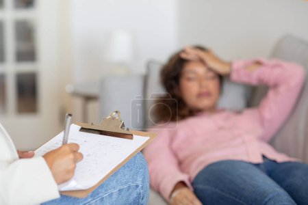 Thérapeute méconnaissable écoutant une cliente allongée sur un canapé à son bureau, prenant des notes au presse-papiers