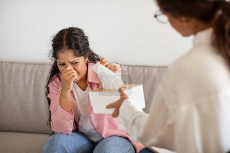 Upset young woman crying during therapy session, sitting on couch in front of lady counselor. Therapist offer tissue to sad client