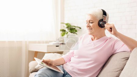 Senior woman is seated on a couch, wearing headphones and listening to music. She appears engaged and focused on the music playing. The room is relaxed and comfortable, with soft lighting.