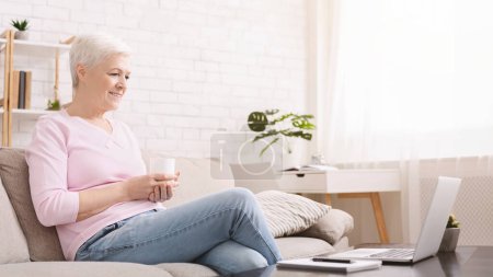 Photo for Senior woman sitting on sofa and watching movie online on laptop, empty space - Royalty Free Image