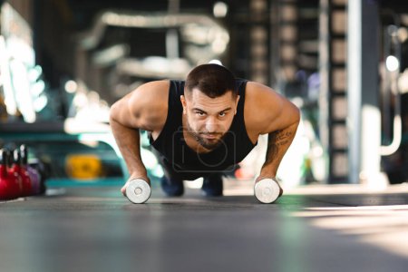 A man is performing push ups while holding two dumbbells in his hands. He is engaging his chest, triceps, and core muscles in this strength training exercise.