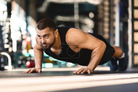 A man is vigorously performing push ups on the floor of a gym while being surrounded by exercise equipment. He shows determination and focus in his workout routine.