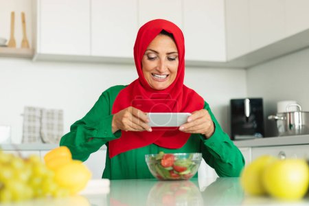 Foto de Una mujer alegre, adornada con un hiyab rojo y un top verde, captura un momento con su smartphone, centrándose en una colorida ensalada colocada en una mesa de cocina - Imagen libre de derechos