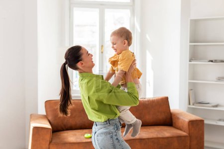 A woman joyfully holds a baby up in the air, both smiling. The kid arms are outstretched, enjoying the playful interaction with the mother at home