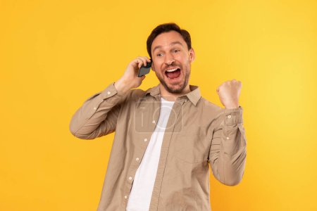 A delighted man is standing against a vibrant yellow backdrop, cheering and making a triumphant fist pump as he converses enthusiastically on his mobile phone, expressing joy or success.