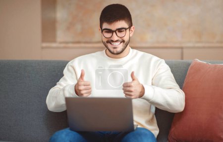 Foto de Un hombre está sentado en un sofá, sosteniendo un portátil en su regazo. Se le ve dando un gesto de pulgar hacia arriba, indicando aprobación o acuerdo con algo en la pantalla. - Imagen libre de derechos