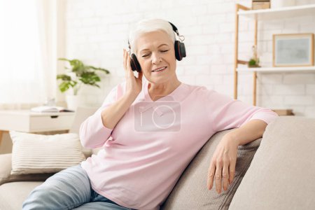 Photo for Senior woman is seated on a couch, wearing headphones and listening intently. She appears relaxed and focused on the music or audio she is enjoying. - Royalty Free Image