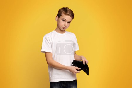 No pocket money. Sad teen boy showing old empty wallet, orange studio background