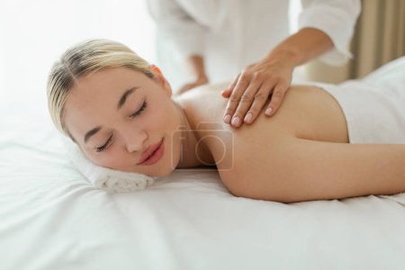 A woman lying face down on a massage table at a luxury spa while a massage therapist is massaging her upper back. The room is dimly lit with soothing music playing in the background.