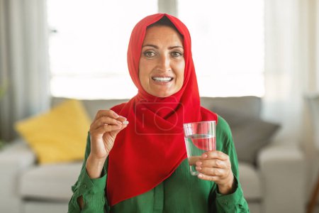 A cheerful woman with her head covered in a vibrant red hijab is holding a pill and a clear glass of water, seemingly preparing to take the medication as she stands inside a well-lit room.