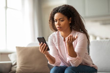 Foto de Esta imagen muestra a una mujer afroamericana en su sofá, frunciendo el ceño ante su teléfono, rodeada de un ambiente hogareño cómodo, que representa un momento de estrés. - Imagen libre de derechos