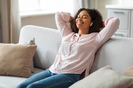 Entspannte junge Afroamerikanerin genießt einen friedlichen Moment auf dem Sofa mit geschlossenen Augen und Kopfhörern und hört Musik