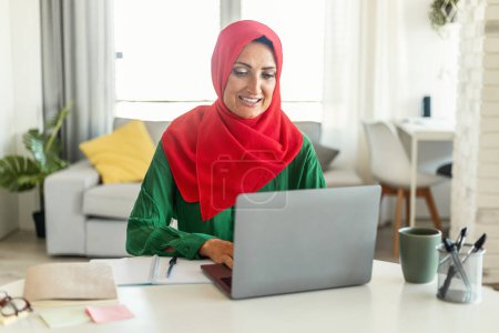 Téléchargez les photos : Femme musulmane assise à une table, concentrée sur l'écran de son ordinateur portable. Elle dactylographie et fait défiler, peut-être travailler ou étudier. Le fond est simple et discret. - en image libre de droit