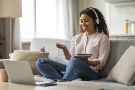 Foto de Joven mujer negra sonriendo, usando auriculares y usando una tableta mientras está sentada cómodamente en su sofá con un portátil a su lado - Imagen libre de derechos