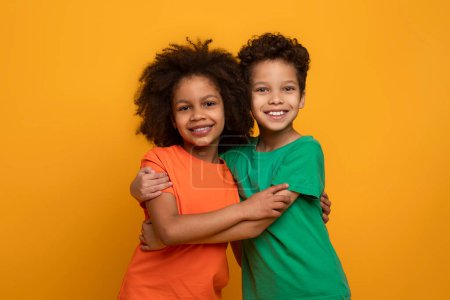 Two young African American children, a boy and a girl, are embracing each other in a warm hug. They stand in front of a bright yellow background, showing affection and closeness.