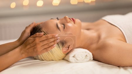Eine Frau liegt in Ruhe auf einem Massagetisch, ihr Gesichtsausdruck ist von tiefer Entspannung geprägt, während geschickte Hände eine beruhigende Kopfmassage durchführen..