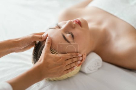 A woman is lying down with her eyes closed, receiving a relaxing facial massage. The therapists hands are gently massaging her face, view above