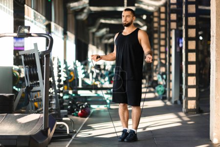 A man stands in a gym, holding a skipping rope. He is focused on his exercise routine, preparing to start his skipping workout.