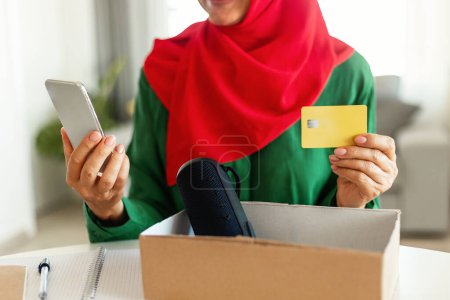 Foto de Una mujer en hiyab está sentada en una mesa, sosteniendo un teléfono inteligente en una mano y una tarjeta de crédito en la otra. Ella parece estar comprometida con el dispositivo, haciendo una compra o comprobando sus finanzas, recortado - Imagen libre de derechos