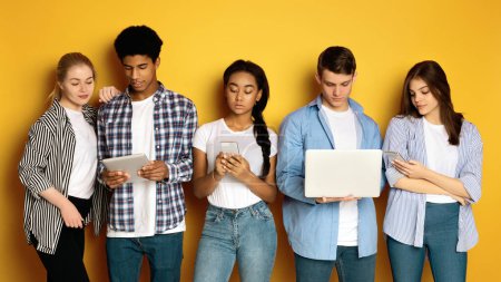 Five multiethnic teenagers stand side by side, each absorbed in different electronic devices, posing on yellow studio background