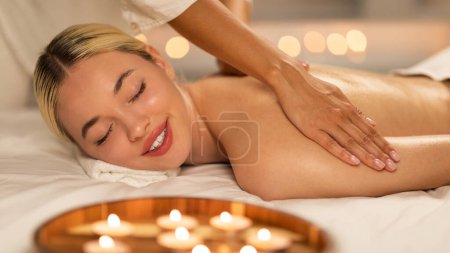 Téléchargez les photos : Heureuse femme blonde s'allonge face contre terre sur une table de massage tandis qu'un thérapeute professionnel pétrit et masse ses muscles du dos pour libérer la tension et favoriser la relaxation. - en image libre de droit