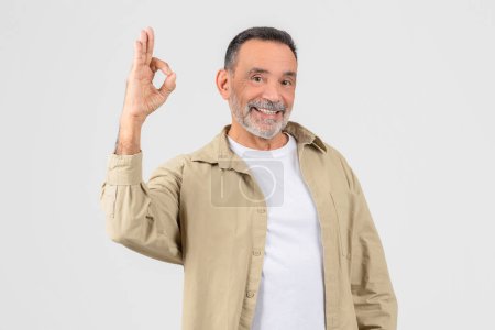 A senior man wearing a white shirt and tan jacket is seen making the Okay sign with his hand. His fingers are split in the iconic OK shape