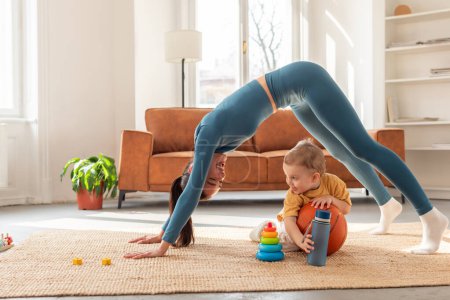 Téléchargez les photos : Une jeune mère est capturée dans une pose de yoga de chien vers le bas, s'étendant sur son bébé ludique qui est assis sur un tapis tissé avec des jouets autour de lui, intérieur de la maison - en image libre de droit
