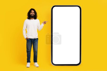 A smiling Indian man with long hair and a beard stands confidently on a bright yellow backdrop, gesturing towards an oversized blank screen of a smartphone model, mockup copy space