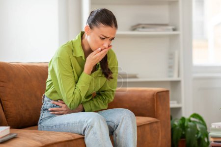 Eine Frau in grünem Hemd und Jeans sitzt auf einem braunen Sofa, blickt verstört mit der Hand über den Mund und umklammert ihren Bauch, ein mögliches Zeichen für Übelkeit oder Bauchschmerzen.