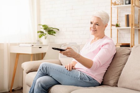 Téléchargez les photos : Femme âgée est assise sur un canapé à l'intérieur, tenant une télécommande dans sa main. Elle semble concentrée sur l'appareil, peut-être regarder la télévision ou ajuster les paramètres - en image libre de droit