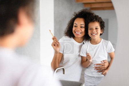 Photo for African American woman and a child standing in front of a bathroom mirror, brushing their teeth with toothbrushes and toothpaste - Royalty Free Image