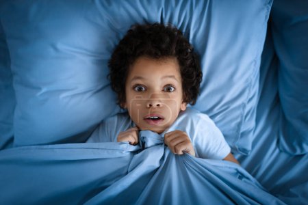 El joven afroamericano yace en la cama con una mirada sorprendida en su cara, agarrándose a la cama azul que cubre su barbilla. Sus amplios ojos transmiten una sensación de asombro o alarma, vista superior