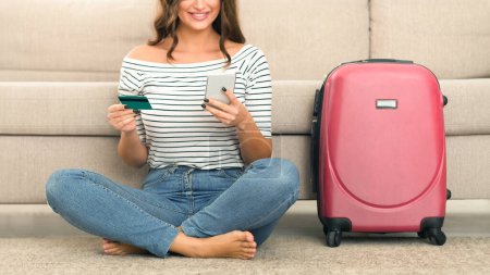 Cropped of woman in casual attire sits on the floor by her couch, holding a credit card and using her smartphone, possibly to book a trip or handle travel arrangements, with a pink suitcase