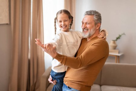 Senior man is standing, holding a little girl in his arms, grandfather dancing with his cute little granddaughter at home