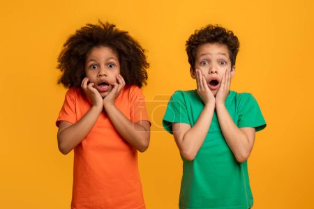Zwei junge afroamerikanische Kinder stehen Seite an Seite, die Augen weit aufgerissen und die Hände auf den Wangen in einem klassischen Ausdruck der Überraschung und des Staunens. Der leuchtend gelbe Hintergrund