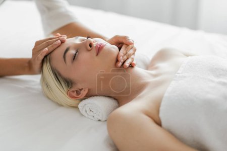 Cette image capture une jeune femme blonde jouissant paisiblement d'un massage facial dans un spa de luxe, mettant en évidence ses expressions faciales détendues