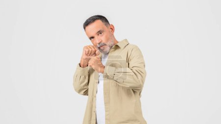 Ein älterer Mann mit braunem Hemd steht da, während er die Hände vor sich in Kampfpose zusammenhält. Er wirkt fokussiert und nachdenklich, als er die Hände zusammenhält..