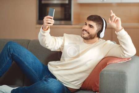 Photo for A cheerful young man reclines comfortably on a sofa wearing casual clothes and wireless headphones. He is holding a smartphone in one hand, have video call with friends - Royalty Free Image