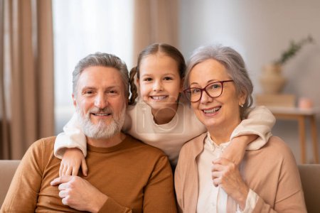 Älterer Mann, Frau und Kind sitzen gemütlich auf einer Couch im Wohnzimmer. Großeltern und Enkelin wirken entspannt und unterhalten sich, machen Fotos