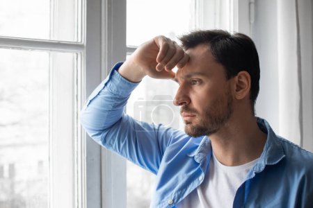 Un hombre está de pie junto a una ventana, mirando afuera. Su mano descansa sobre su cabeza mientras parece perdido en el pensamiento, tal vez contemplando o reflexionando
