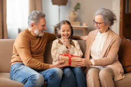 A young girl is seated on a couch between her smiling grandparents, receiving a wrapped gift with a red ribbon. The warmth of the scene is enhanced by the soft lighting and comfortable interior