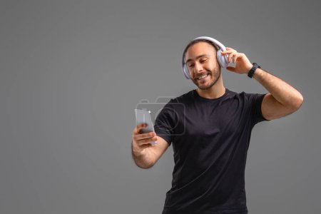 Foto de Un hombre con auriculares sosteniendo un teléfono celular en la mano, dedicado a escuchar música o una llamada telefónica. El atuendo de los hombres y los alrededores sugieren que es probable que en un entorno público, espacio de copia - Imagen libre de derechos