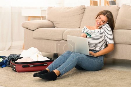 A young woman sitting on the floor of her living room, leaning against a sofa as she converses on her smartphone with a pleased smile, holding credit card and laptop, booking tickets or hotel