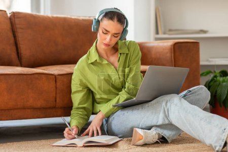 Eine fokussierte junge Frau sitzt mit dem Rücken auf einer Couch in einem gut beleuchteten Raum und hört Musik über Kopfhörer, während sie in ein Notizbuch schreibt, mit offenem Laptop vor sich.