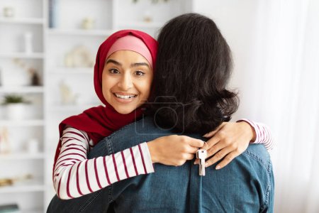 Foto de Una joven oriental que lleva un hiyab rojo está abrazando a su marido por detrás, sosteniendo la llave de su nueva casa. - Imagen libre de derechos